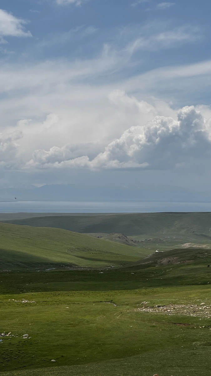 Green rolling pasture in Suusamyr Valley with a lake and mountains in the distance under a cloudy sky